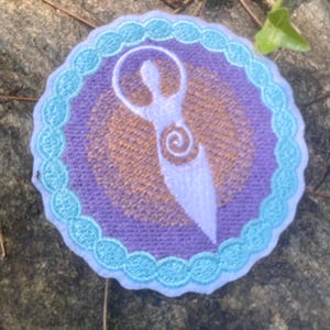 May include: Circular embroidered patch with a white figure and spiral design on a purple background. It has a light blue scalloped border and a golden-brown inner circle. The patch is circular in shape.