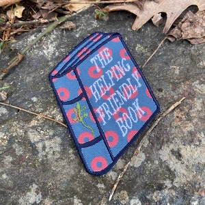 K&ouml;nnte beinhalten: Gestickter Aufn&auml;her in Form eines Buches mit dem Text "THE HELPING FRIENDLY BOOK". Der Aufn&auml;her hat einen blauen Hintergrund mit roten donutartigen Mustern. Auf der Seite des Buches ist eine gr&uuml;ne Eidechse gestickt.