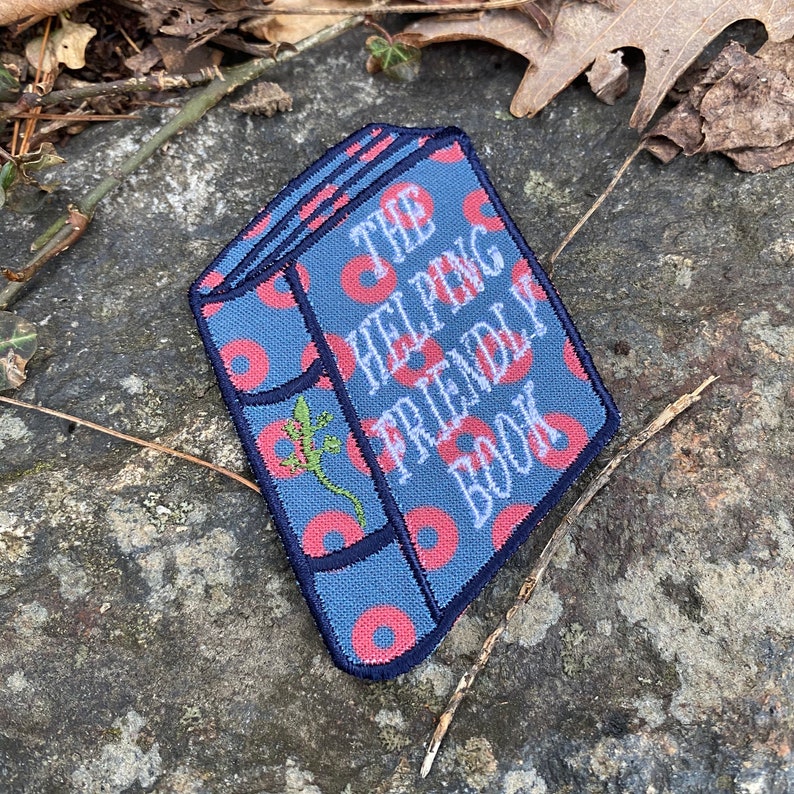 K&ouml;nnte beinhalten: Gestickter Aufn&auml;her in Form eines Buches mit dem Text "THE HELPING FRIENDLY BOOK". Der Aufn&auml;her hat einen blauen Hintergrund mit roten donutartigen Mustern. Auf der Seite des Buches ist eine gr&uuml;ne Eidechse gestickt.