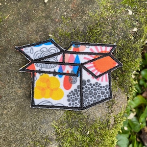 May include: An open box patch with a black outline. The box is filled with colorful patterns, including yellow flowers, orange suns, and blue triangles. The patch is on a mossy rock.
