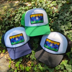 May include: Three trucker hats with a white front panel and a mesh back. Each hat features a rectangular patch with a geometric design in blue, yellow, and green, and a white star. The hats have different colored brims: green, navy, and black.