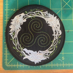 May include: Round black patch with embroidered white wolf heads and a green Celtic knot design. The wolves are framed by green vines. The patch is approximately 4 inches in diameter.
