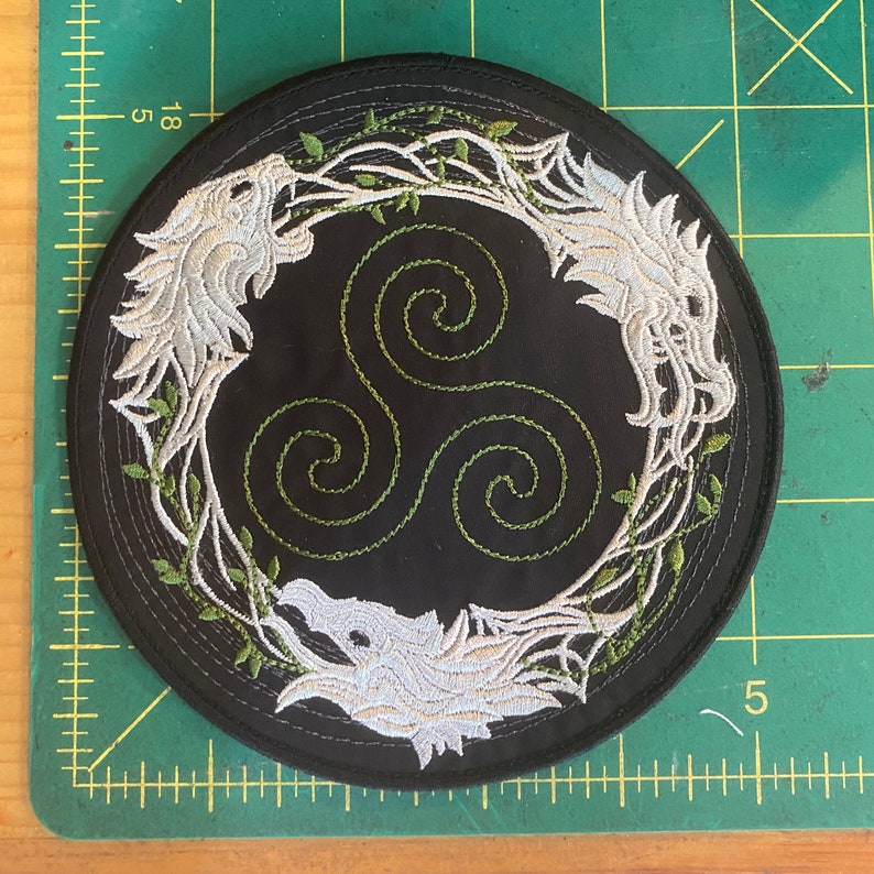 May include: Round black patch with embroidered white wolf heads and a green Celtic knot design. The wolves are framed by green vines. The patch is approximately 4 inches in diameter.