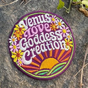 May include: A circular embroidered patch with the text "Venus Love Goddess Creation" in white and pink. The patch has a purple base, a sunrise design, and colourful flowers. It is edged with green.