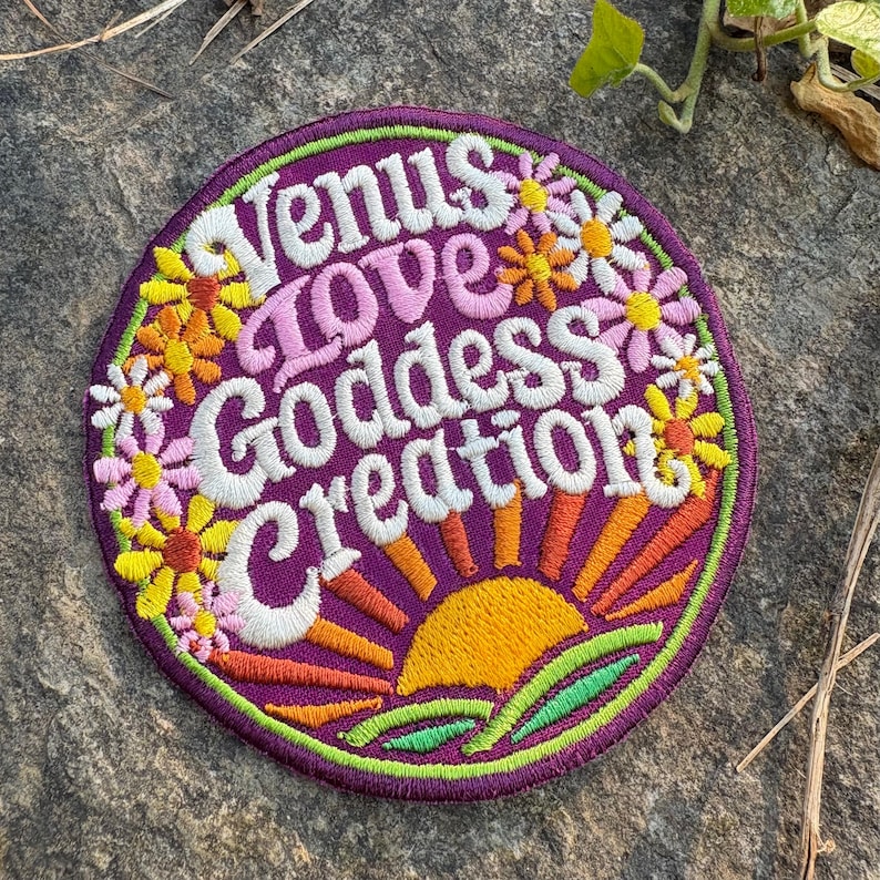 May include: A circular embroidered patch with the text "Venus Love Goddess Creation" in white and pink. The patch has a purple base, a sunrise design, and colourful flowers. It is edged with green.