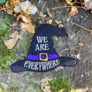 May include: Black witch hat patch with the words "WE ARE EVERYWHERE" embroidered in white. The hat has a purple band with a gold buckle. The patch is on a textured gray stone surface with moss and dried leaves.