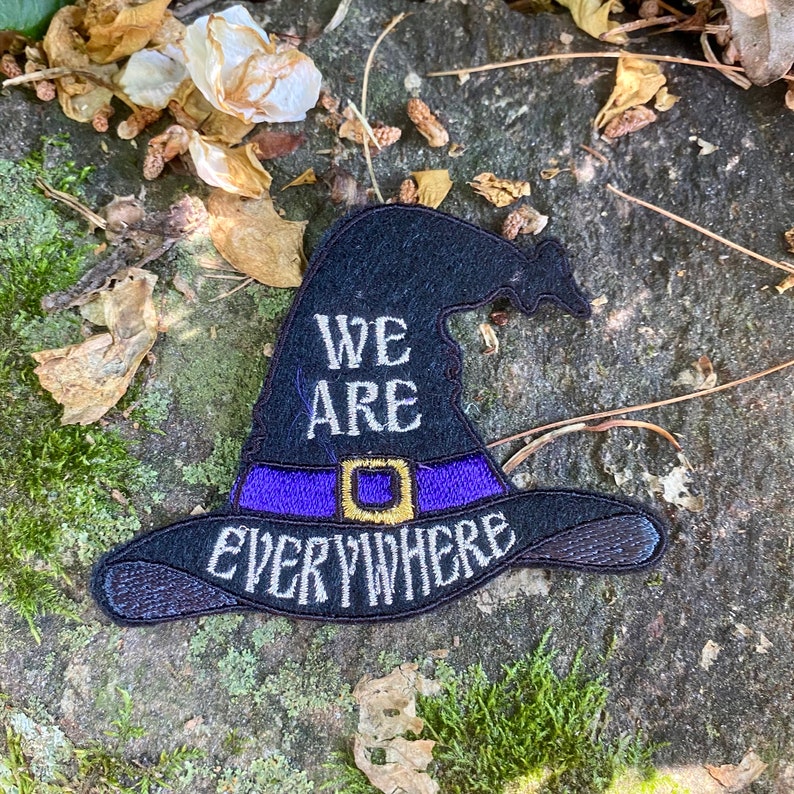 May include: Black witch hat patch with the words "WE ARE EVERYWHERE" embroidered in white. The hat has a purple band with a gold buckle. The patch is on a textured gray stone surface with moss and dried leaves.