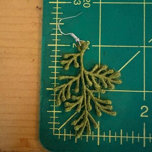 May include: A single olive green embroidered earring in a fern-like design. The earring has a silver-colored hook. The earring is on a green cutting mat with inch markings.