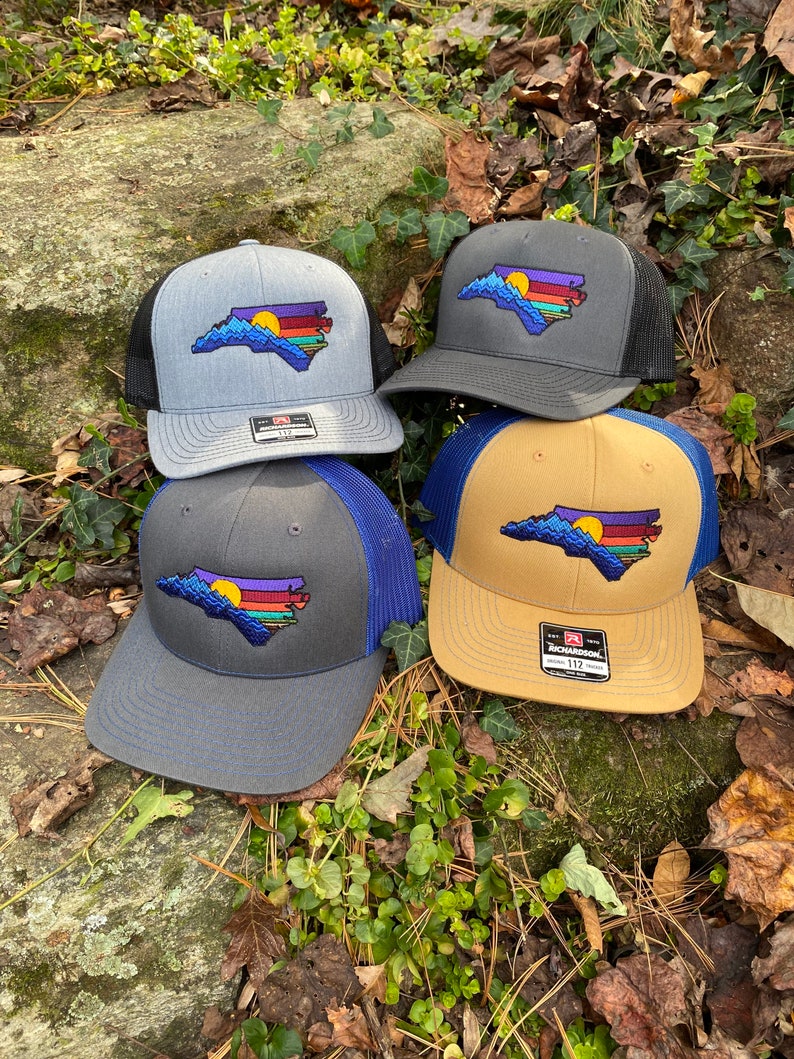 Pode incluir: Quatro bon&eacute;s de caminhoneiro com um contorno bordado do estado da Carolina do Norte com um desenho de p&ocirc;r do sol. Os bon&eacute;s t&ecirc;m diferentes combina&ccedil;&otilde;es de cores: cinza com malha preta, cinza escuro com malha preta, cinza com malha azul e bege com malha azul.