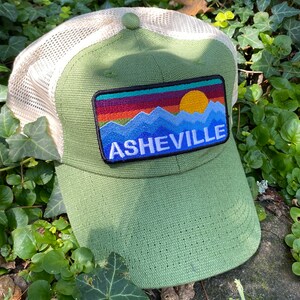 Peut inclure: Casquette de baseball verte avec un dos en filet et un &eacute;cusson repr&eacute;sentant un paysage de montagne et le mot "ASHEVILLE" en lettres blanches. L'&eacute;cusson comprend un coucher de soleil aux teintes orange, rouge et jaune.