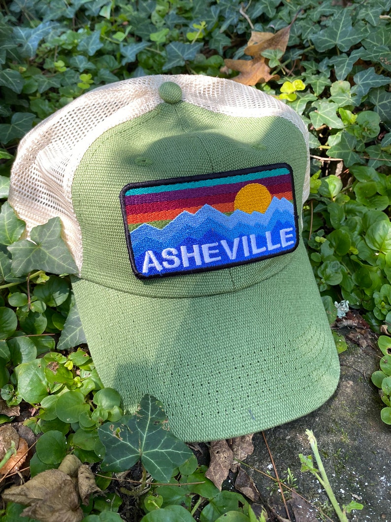 Peut inclure: Casquette de baseball verte avec un dos en filet et un &eacute;cusson repr&eacute;sentant un paysage de montagne et le mot "ASHEVILLE" en lettres blanches. L'&eacute;cusson comprend un coucher de soleil aux teintes orange, rouge et jaune.