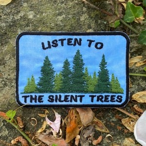 May include: Embroidered patch with a light blue background featuring a forest scene with green trees and the text "LISTEN TO THE SILENT TREES" in black. The patch has a black border.
