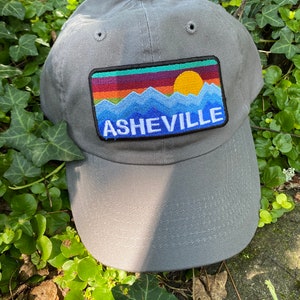 Peut inclure: Casquette de baseball grise avec un &eacute;cusson rectangulaire repr&eacute;sentant un paysage de montagne et de coucher de soleil. L'&eacute;cusson pr&eacute;sente une cha&icirc;ne de montagnes bleue, un soleil jaune et le mot "ASHEVILLE" en bleu. La casquette est en tissu doux.