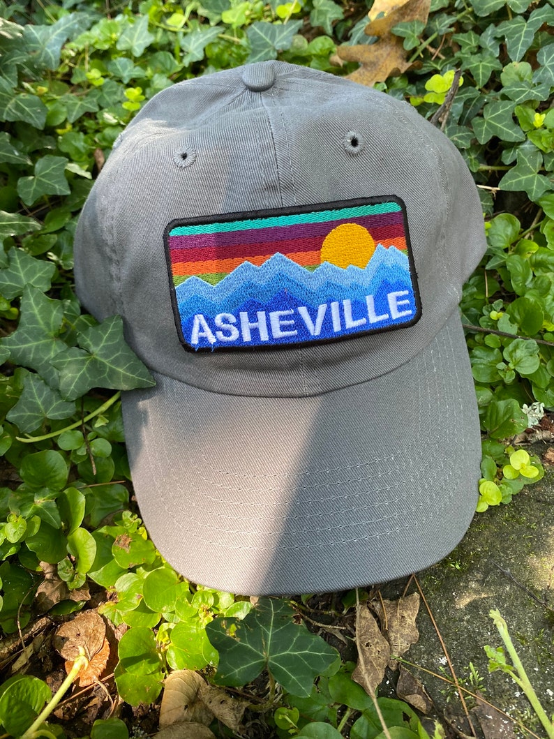Peut inclure: Casquette de baseball grise avec un &eacute;cusson rectangulaire repr&eacute;sentant un paysage de montagne et de coucher de soleil. L'&eacute;cusson pr&eacute;sente une cha&icirc;ne de montagnes bleue, un soleil jaune et le mot "ASHEVILLE" en bleu. La casquette est en tissu doux.