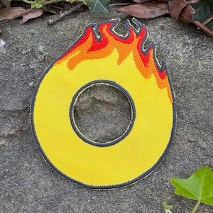 Puede incluir: Parche bordado amarillo con forma de cero y dise&ntilde;o de llama. Las llamas son rojas y naranjas, y el parche tiene un borde gris oscuro. El parche est&aacute; sobre una superficie de piedra gris.