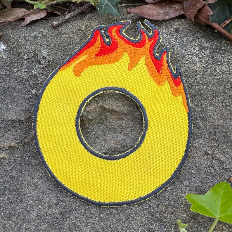 Puede incluir: Parche bordado amarillo con forma de cero y dise&ntilde;o de llama. Las llamas son rojas y naranjas, y el parche tiene un borde gris oscuro. El parche est&aacute; sobre una superficie de piedra gris.