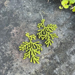 May include: A pair of lime green leaf-shaped earrings. The earrings are made of a textured material and feature intricate details resembling foliage. The earrings hang from silver-colored hooks. The background is a mottled gray surface.