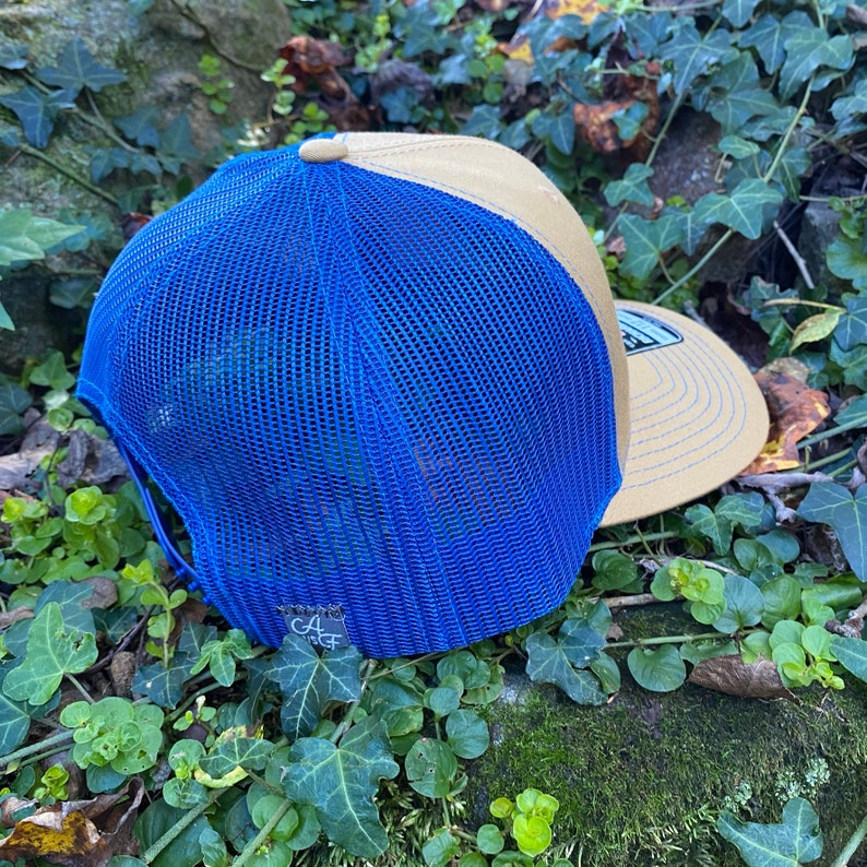 Pode incluir: Bon&eacute; de caminhoneiro azul e bege com costas em malha. O bon&eacute; tem uma aba curva e um patch com as letras "CA" e um pequeno desenho. O bon&eacute; est&aacute; sobre um leito de folhas verdes e musgo.