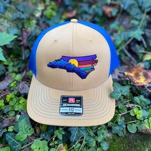Pode incluir: Bon&eacute; de caminhoneiro bege e azul com um design bordado do contorno do estado da Carolina do Norte, retratando montanhas e um p&ocirc;r do sol. O bon&eacute; tem uma aba curva e uma parte traseira de malha. A etiqueta da marca Richardson &eacute; vis&iacute;vel.