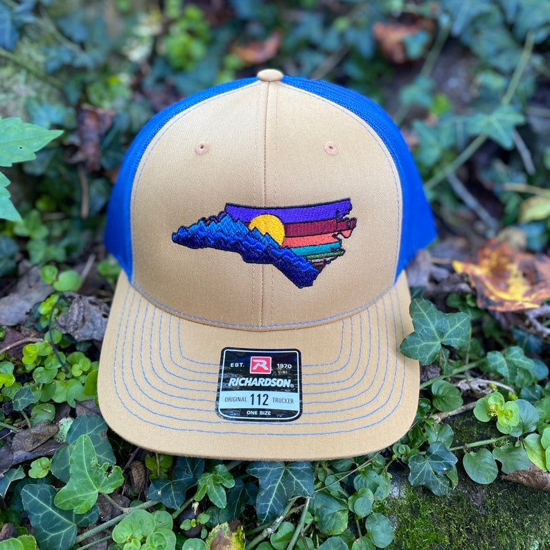 Pode incluir: Bon&eacute; de caminhoneiro bege e azul com um design bordado do contorno do estado da Carolina do Norte, retratando montanhas e um p&ocirc;r do sol. O bon&eacute; tem uma aba curva e uma parte traseira de malha. A etiqueta da marca Richardson &eacute; vis&iacute;vel.