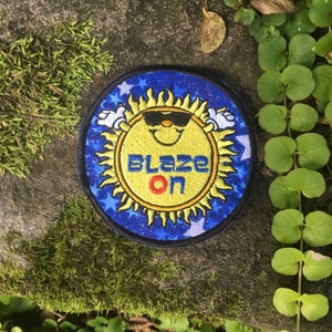 Puede incluir: Parche redondo con un sol sonriente con gafas de sol, rodeado de llamas amarillas y manos blancas. El parche tiene un fondo azul con estrellas blancas. Las palabras "BLAZE ON" est&aacute;n en el centro, con "BLAZE" en azul y "ON" en rojo.