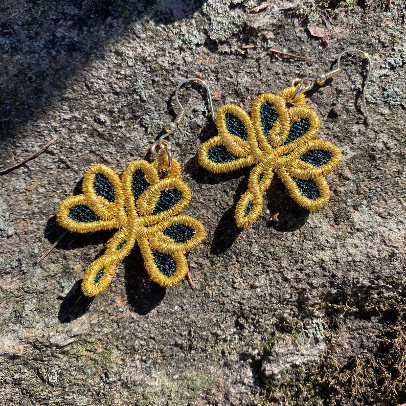 Pode incluir: Um par de brincos bordados dourados com detalhes em preto em forma de lib&eacute;lula. Os brincos t&ecirc;m um design delicado e vazado e s&atilde;o suspensos por ganchos prateados. S&atilde;o exibidos numa superf&iacute;cie texturizada cinza-castanha.