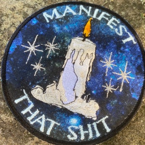 May include: Round patch with a blue and black galaxy background. A white candle with a yellow flame is centered, with the words "MANIFEST THAT SHIT" in white. Silver starbursts are scattered around the candle.