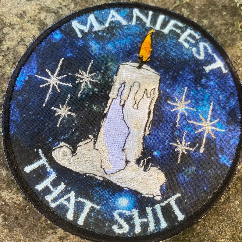 May include: Round patch with a blue and black galaxy background. A white candle with a yellow flame is centered, with the words "MANIFEST THAT SHIT" in white. Silver starbursts are scattered around the candle.