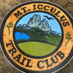 Op de afbeelding: Ronde patch met de tekst "MT. ICCULUS TRAIL CLUB" in zwarte letters. De patch toont een bergketen, een meer en een vogel tegen een blauwe lucht. De rand is oranje met groene bladaccenten.