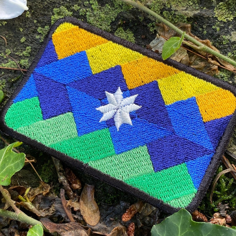 May include: Embroidered patch with a geometric design in shades of blue, green, and yellow, featuring a white star in the center. The patch has a black border and is rectangular in shape. The patch is likely for clothing or accessories.