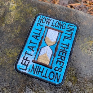 May include: Embroidered patch with a blue background and a black border. The patch features an hourglass design with the text "HOW LONG UNTIL THERE'S NOTHING LEFT AT ALL?" in white lettering. The hourglass has tan sand.