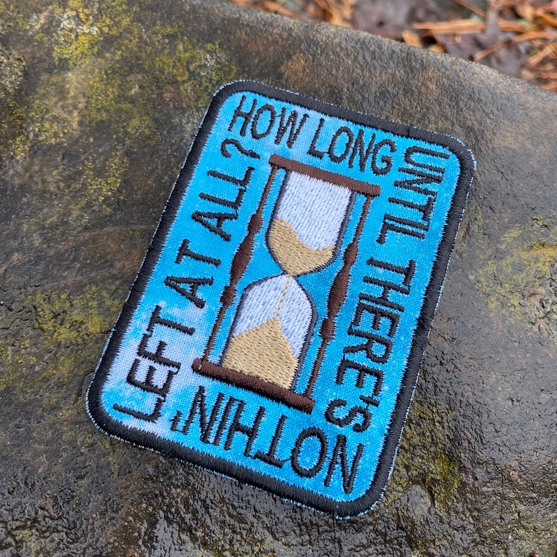 May include: Embroidered patch with a blue background and a black border. The patch features an hourglass design with the text "HOW LONG UNTIL THERE'S NOTHING LEFT AT ALL?" in white lettering. The hourglass has tan sand.