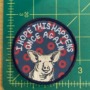May include: Round patch with a navy blue background featuring red circles and a pig illustration. The text "I HOPE THIS HAPPENS ONCE AGAIN" is embroidered in white. The patch has a dark blue border.