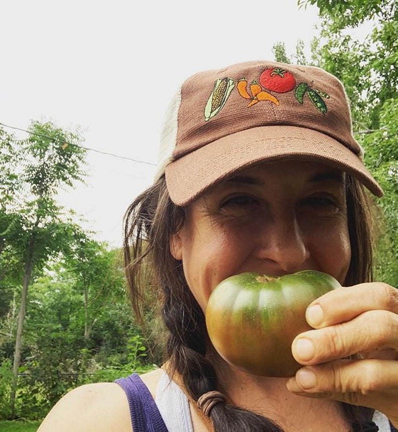 May include: A brown trucker hat with a white mesh back and embroidered vegetables, including corn, peppers, a tomato, and peas. A person is holding a large green tomato. The background features trees and a cloudy sky.