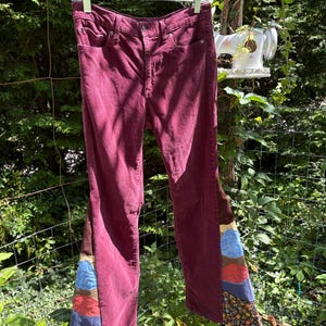 Op de afbeelding: Bordeaux corduroy broek met patchwork flared pijpen. Het patchwork heeft bruine, blauwe en bloemmotief stoffen. De broek hangt buiten aan een doorzichtige plastic hanger.