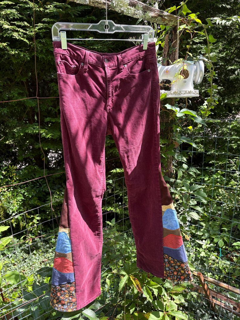 Op de afbeelding: Bordeaux corduroy broek met patchwork flared pijpen. Het patchwork heeft bruine, blauwe en bloemmotief stoffen. De broek hangt buiten aan een doorzichtige plastic hanger.