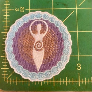 May include: Embroidered patch with a white stylized figure with arms raised, set within a purple circle with a gold background. The circle has a light blue scalloped edge. The patch is on a green cutting mat.