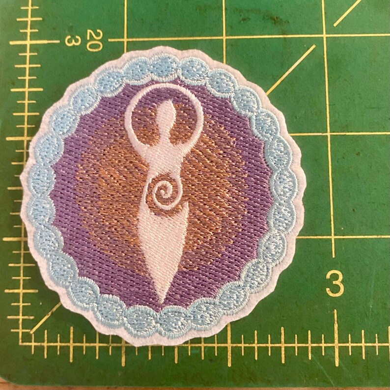May include: Embroidered patch with a white stylized figure with arms raised, set within a purple circle with a gold background. The circle has a light blue scalloped edge. The patch is on a green cutting mat.