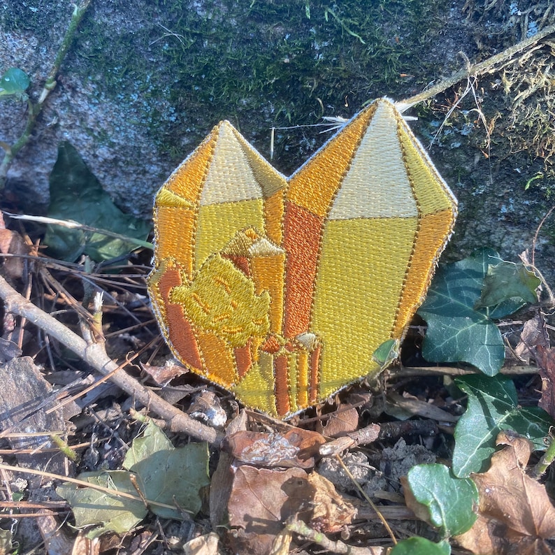May include: Embroidered patch with a crystal design in yellow, orange, and cream hues. The patch has a textured look with detailed stitching. It is set against a natural background of leaves and twigs.