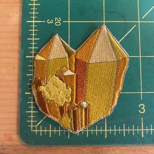 May include: Embroidered patch in the shape of a heart-shaped crystal formation. The patch features shades of gold, orange, and brown, with a detailed, textured design. The patch is approximately 7.6 cm wide.