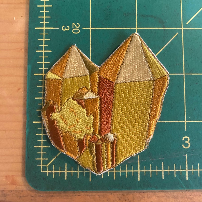 May include: Embroidered patch in the shape of a heart-shaped crystal formation. The patch features shades of gold, orange, and brown, with a detailed, textured design. The patch is approximately 7.6 cm wide.
