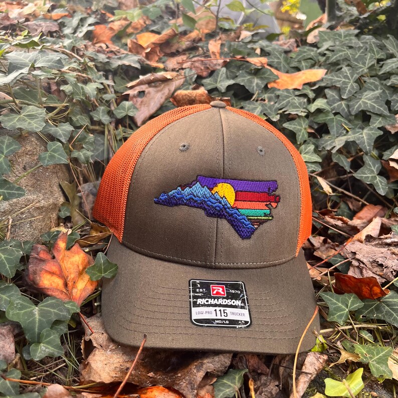 K&ouml;nnte beinhalten: Braune und orangefarbene Trucker-Cap mit gesticktem Design des Umrisses des Bundesstaates North Carolina. Das Design umfasst eine Bergkette, eine Sonne und farbige Streifen. Die Kappe hat einen gebogenen Schirm.