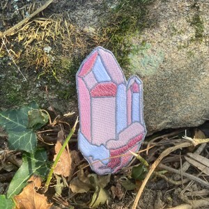May include: Embroidered patch in the shape of a crystal geode. The patch features shades of pink, red, and blue, with a grey outline. The patch is placed on a stone surface with leaves and twigs.