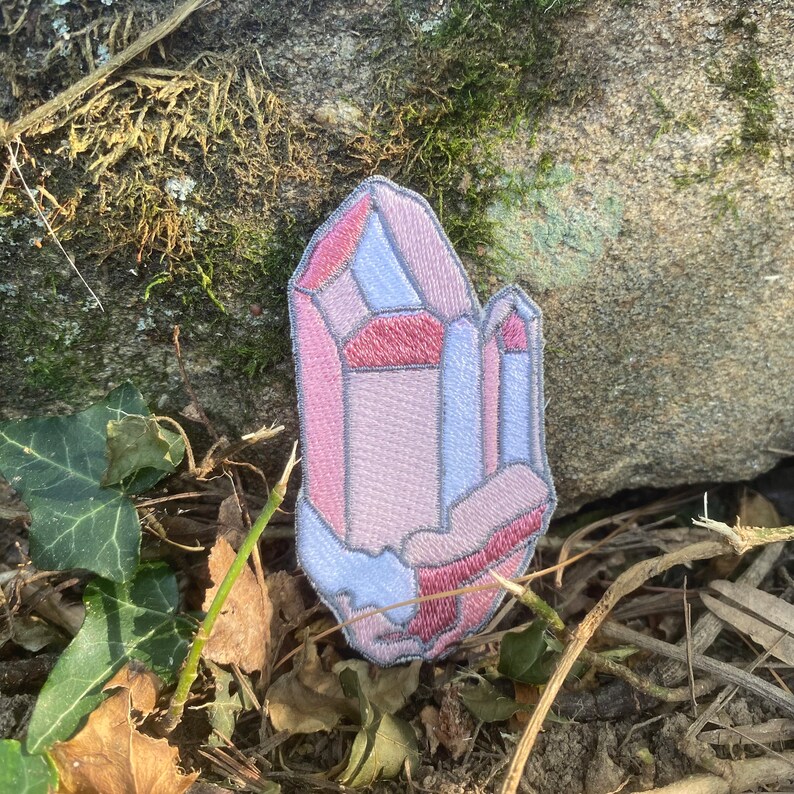 May include: Embroidered patch in the shape of a crystal geode. The patch features shades of pink, red, and blue, with a grey outline. The patch is placed on a stone surface with leaves and twigs.
