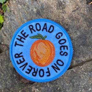 May include: A round, blue embroidered patch with the text "THE ROAD GOES ON FOREVER" in black. The patch features an orange peach with green leaves in the center. The patch is on a gray stone surface.