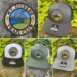 Op de afbeelding: Assortiment van Richardson 112 trucker caps in diverse kleuren, elk met een cirkelvormige patch met de tekst "MT. ICCULUS TRAIL CLUB". De patch toont een berglandschap met een meer en blauwe lucht. Kleuren zijn grijs, wit en groen.