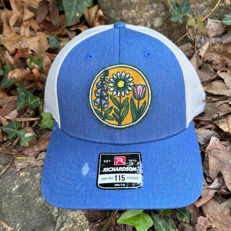 May include: Blue trucker cap with a white mesh back and a floral patch. The patch features a daisy, pink tulip, and other flowers. The cap has a curved peak and a tag that reads "RICHARDSON 115 TRUCKER".