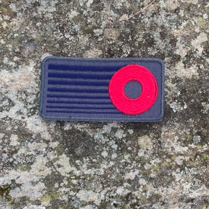 K&ouml;nnte beinhalten: Ein rechteckiger Aufn&auml;her mit dunkelblauem Hintergrund und horizontalen Linien, mit einem roten Kreis mit einem Loch in der Mitte. Der Aufn&auml;her hat einen grauen Rand, wahrscheinlich ein Emblem oder Abzeichen.