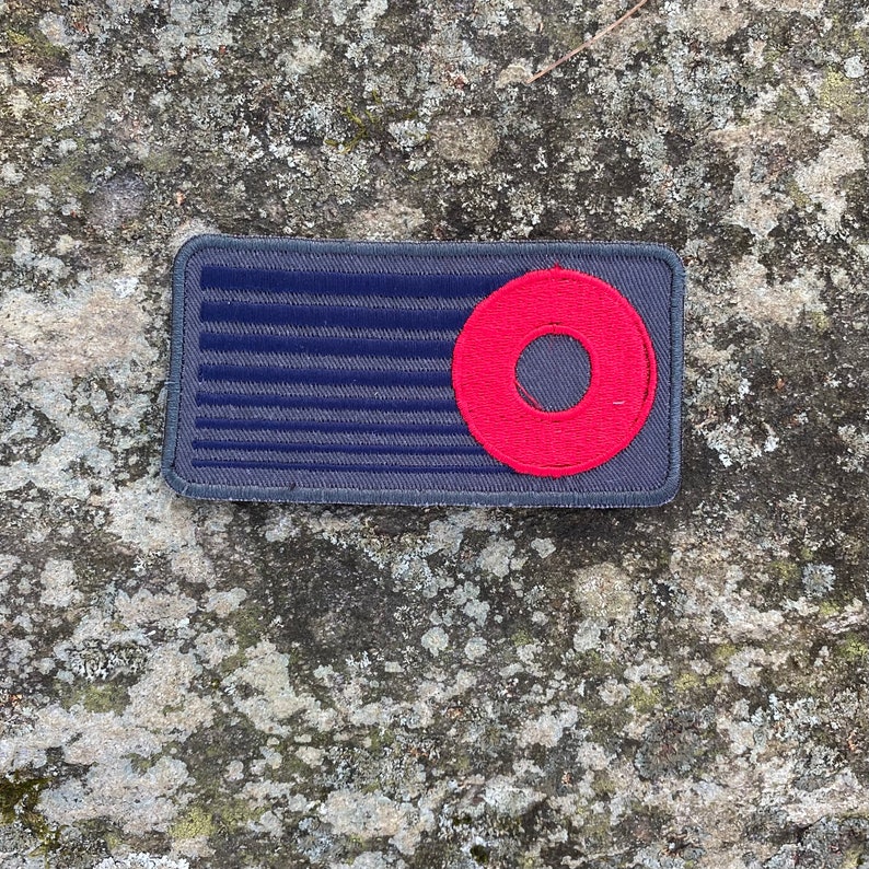 K&ouml;nnte beinhalten: Ein rechteckiger Aufn&auml;her mit dunkelblauem Hintergrund und horizontalen Linien, mit einem roten Kreis mit einem Loch in der Mitte. Der Aufn&auml;her hat einen grauen Rand, wahrscheinlich ein Emblem oder Abzeichen.