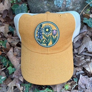 May include: Mustard yellow baseball cap with a white mesh back. A circular patch on the front features a floral design with a daisy, purple flowers, and a pink tulip. The hat is sitting on a bed of leaves.